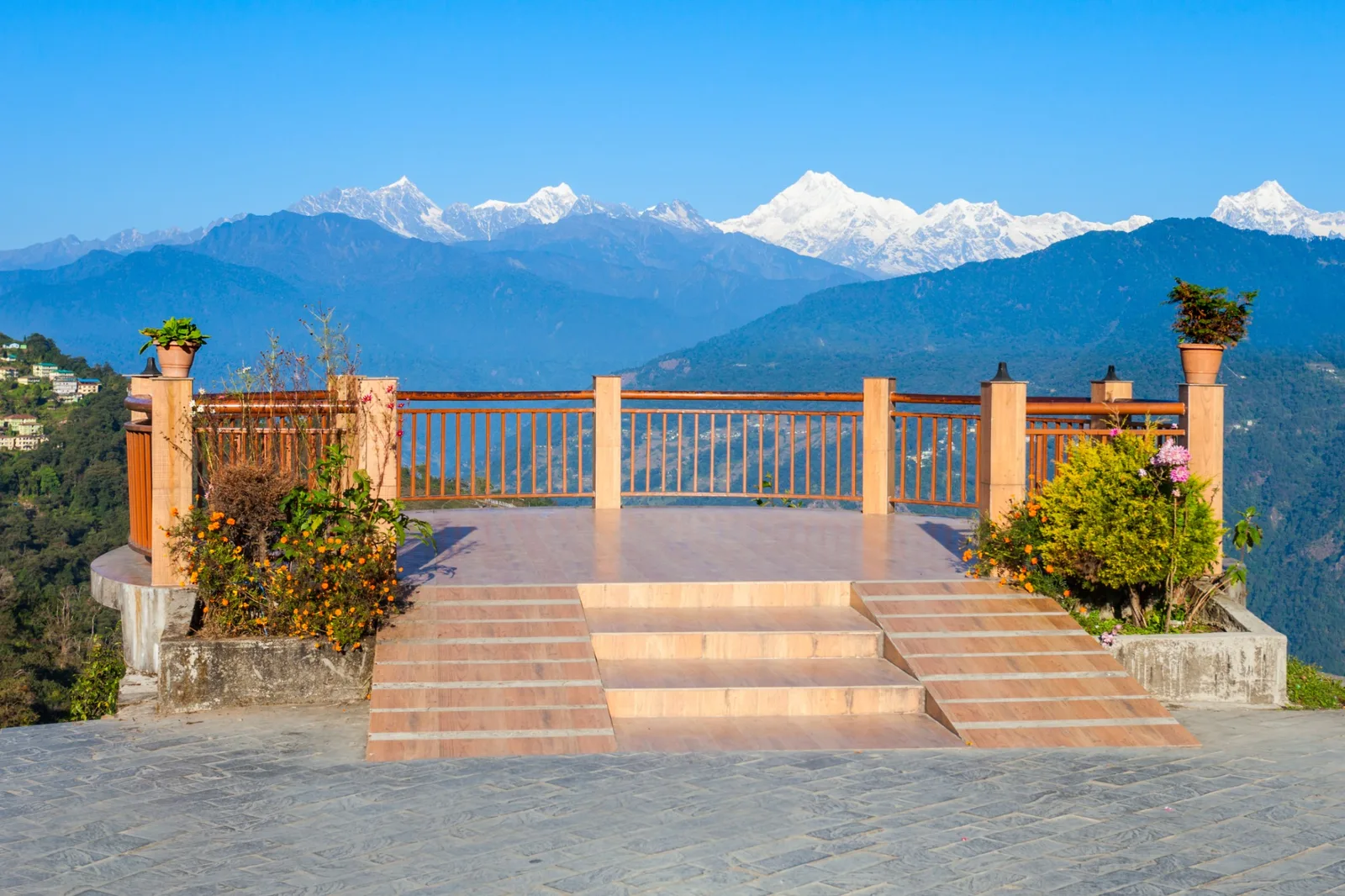 A tiled viewing deck with a wooden railing and flowerpots, looking out over forested hills to a long range of snow peaks under a clear blue sky