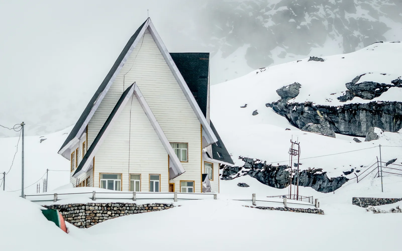 A white wooden building with a steep roof in deep snow, below rocky snow-covered slopes