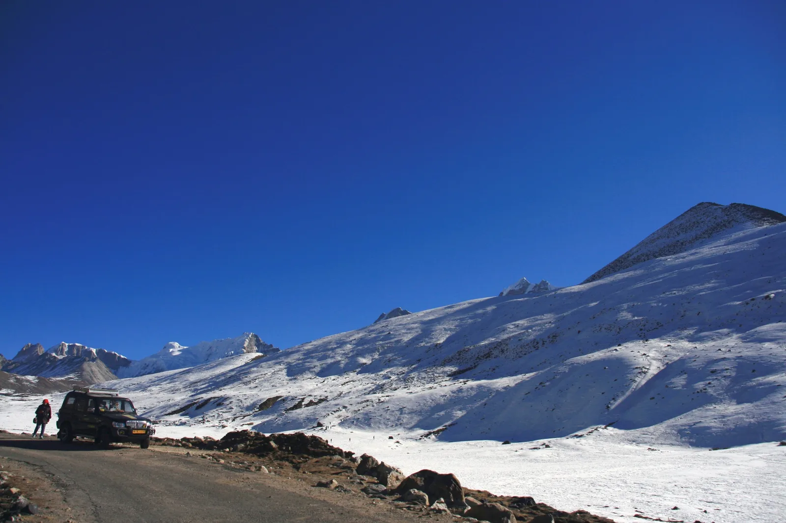 A dark four-wheel-drive car parked on a mountain road beside wide snow-covered slopes and peaks, a traveller in a red jacket next to it, under a deep blue sky